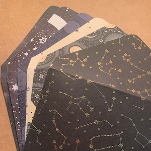 Puede incluir: Una pila de sobres negros con constelaciones y estrellas doradas en lámina. Los sobres son perfectos para enviar cartas o invitaciones.