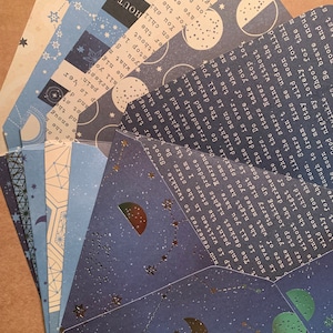 May include: A set of six folded cards with a celestial theme. The cards feature a dark blue background with silver stars and moons, and the text "The darkest nights produce the brightest stars." The cards are perfect for adding a touch of whimsy to your home decor or for sending a special message to someone you love.