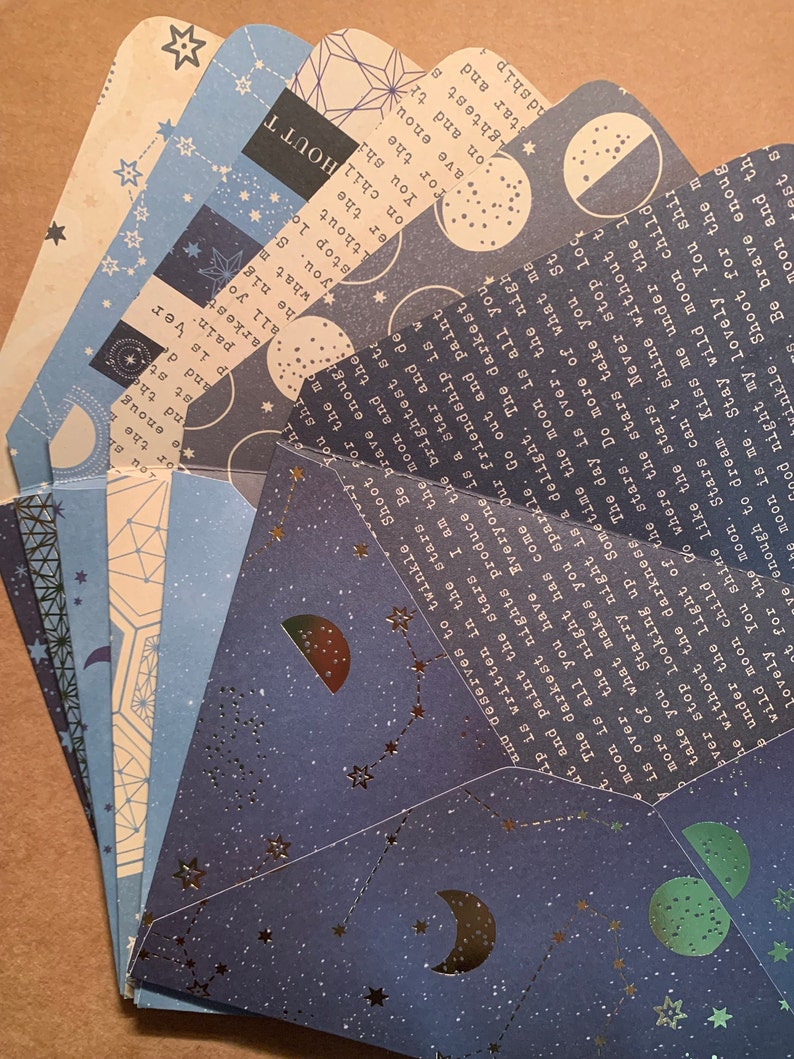 May include: A set of six folded cards with a celestial theme. The cards feature a dark blue background with silver stars and moons, and the text "The darkest nights produce the brightest stars." The cards are perfect for adding a touch of whimsy to your home decor or for sending a special message to someone you love.