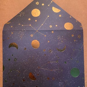 May include: A dark blue envelope with a gold foil design of stars, moons, and constellations. The envelope has a subtle glitter finish.