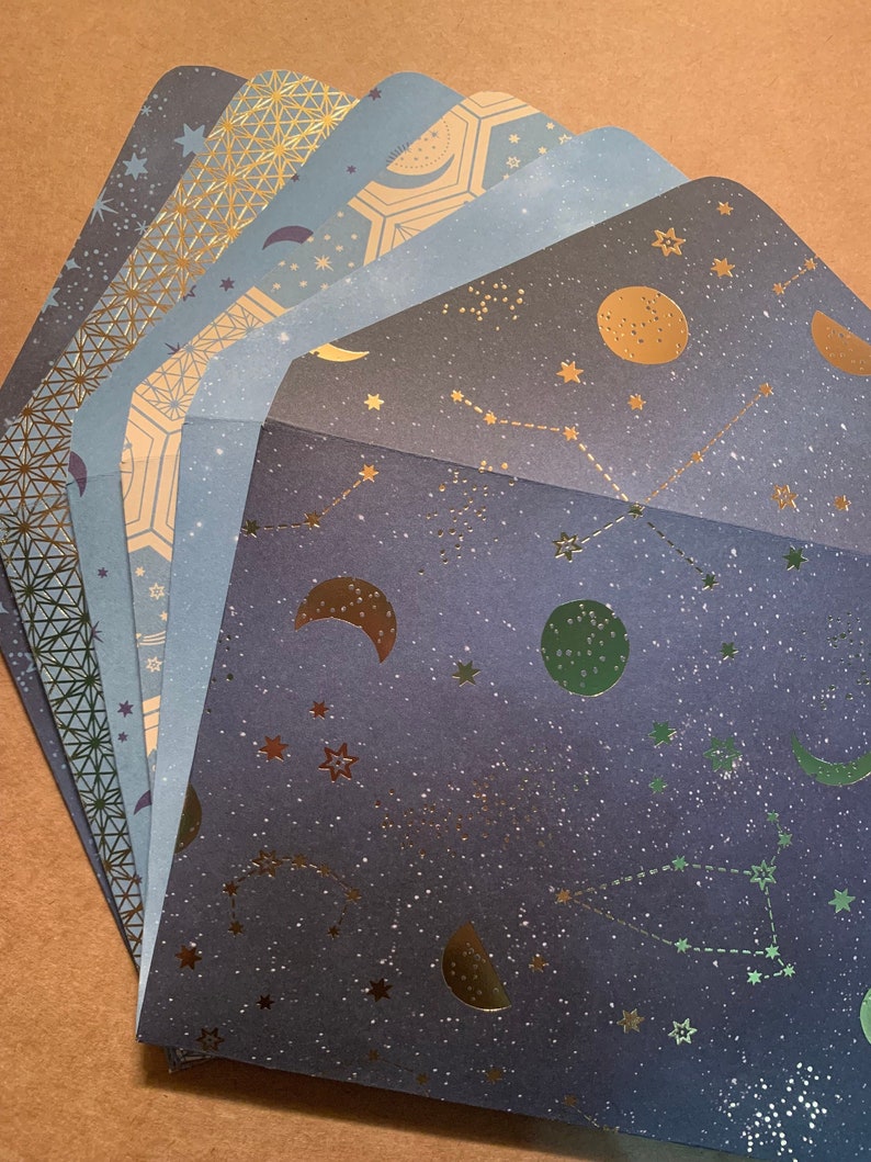 May include: A stack of six blue and gold patterned envelopes with a celestial theme. The envelopes feature stars, moons, and constellations.