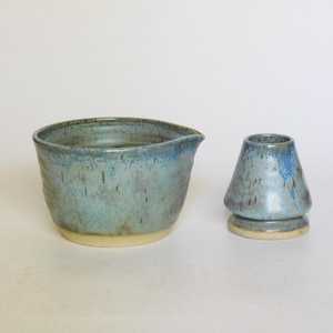 May include: A ceramic matcha bowl with a pouring spout and a matching whisk holder. Both pieces feature a variegated blue and grey glaze with brown speckles and a smooth, unglazed beige base. The bowl has a wide, rounded shape, while the whisk holder is conical.