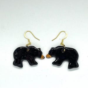 May include: A pair of black bear-shaped earrings with gold-coloured hooks. The bears have small, tan-coloured snouts. The earrings are set against a white background.