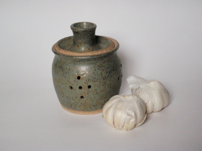 May include: A green ceramic garlic keeper with holes for ventilation. Two white garlic bulbs are in front of the keeper.