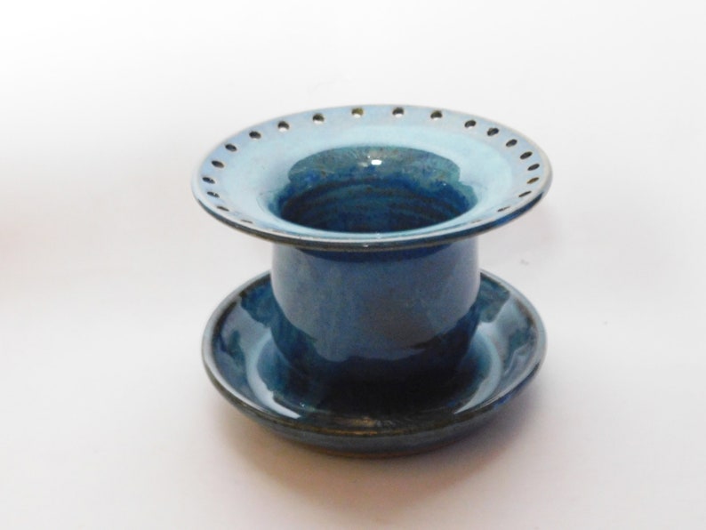 Pu&ograve; includere: Un vaso di fiori in ceramica blu con un sottovaso. Il vaso ha una parte superiore larga e piatta con fori attorno al bordo. Il sottovaso &egrave; dello stesso colore blu.