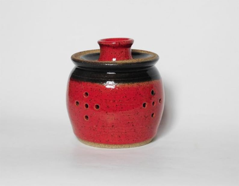 May include: Red and black ceramic jar with holes for storing garlic or onions. The jar has a speckled glaze and a lid.