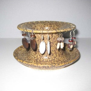 Pu&ograve; includere: Un porta orecchini in ceramica con una glassa macchiata di marrone e beige. Il porta orecchini ha una base rotonda e un centro cilindrico alto con fori per gli orecchini. Ci sono cinque paia di orecchini sul porta orecchini, tra cui orecchini in legno marrone, orecchini in conchiglia bianca e orecchini con perline marroni e bianche.