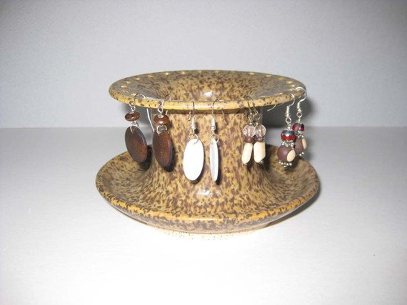 Pu&ograve; includere: Un porta orecchini in ceramica con una glassa macchiata di marrone e beige. Il porta orecchini ha una base rotonda e un centro cilindrico alto con fori per gli orecchini. Ci sono cinque paia di orecchini sul porta orecchini, tra cui orecchini in legno marrone, orecchini in conchiglia bianca e orecchini con perline marroni e bianche.