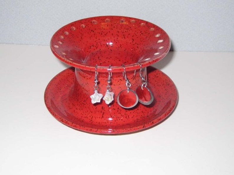 Pu&ograve; includere: Porta orecchini in ceramica rossa con design a due livelli. Il livello superiore ha fori per gli orecchini e il livello inferiore &egrave; un piattino. Ci sono tre paia di orecchini sul supporto.
