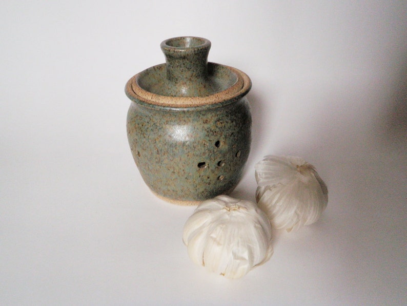 May include: A green ceramic garlic keeper with holes for ventilation. The keeper has a lid with a small opening on top. Two whole garlic bulbs are in front of the keeper.