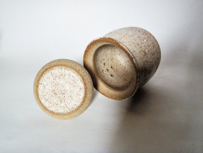 May include: A set of two ceramic salt and pepper shakers with a speckled white glaze. The shakers have a round shape and a small opening on top.