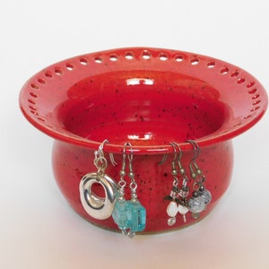 Puede incluir: Porta pendientes de cer&aacute;mica roja con un borde festoneado y agujeros para colgar pendientes. El porta pendientes tiene un esmalte moteado y est&aacute; lleno de pendientes coloridos.