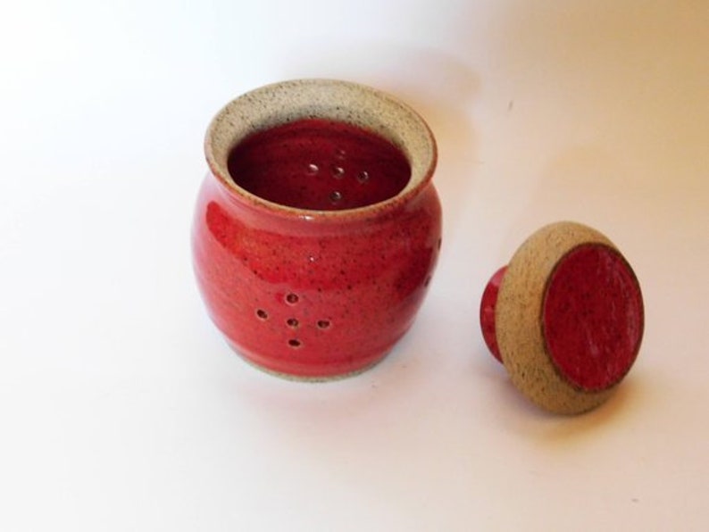 May include: Red ceramic jar with a white lid and holes in the side. The jar is speckled with white and has a red lid.