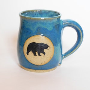 May include: A blue ceramic mug with a white circle and a black bear silhouette. The mug has a light blue glaze with a speckled finish.