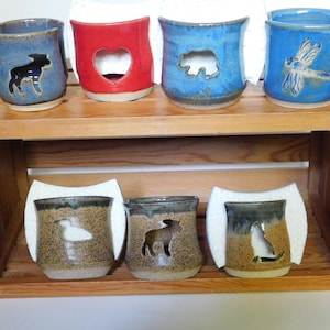 May include: Six ceramic mugs with various animal silhouettes cut out. The mugs are arranged in two rows on a wooden shelf. The top row features a blue mug with a moose, a red mug with a heart, a blue mug with a bear, and a blue mug with a dragonfly. The bottom row features a brown mug with a bird, a brown mug with a moose, and a brown mug with a cat.