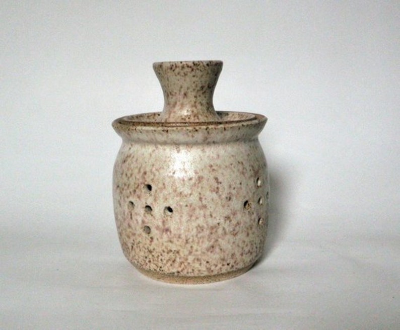 May include: A white ceramic jar with brown speckles and holes. The jar has a lid with a small opening in the center.