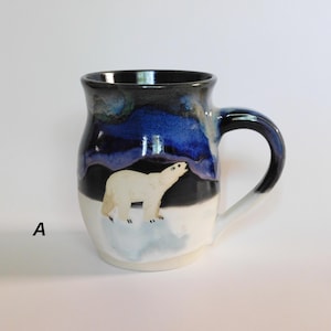 May include: A ceramic mug featuring a hand-painted white polar bear standing on a light blue ice patch. The upper portion of the mug is glazed in deep blue and black, representing a night sky, while the lower section is white. The letter A is visible.