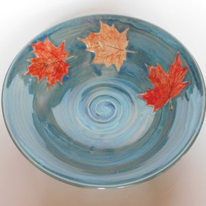 Peut inclure: Un grand bol en céramique bleu avec un motif de glaçure tourbillonnant. Trois feuilles d'érable rouges sont incrustées dans le glaçage, créant un design sur le thème de l'automne.