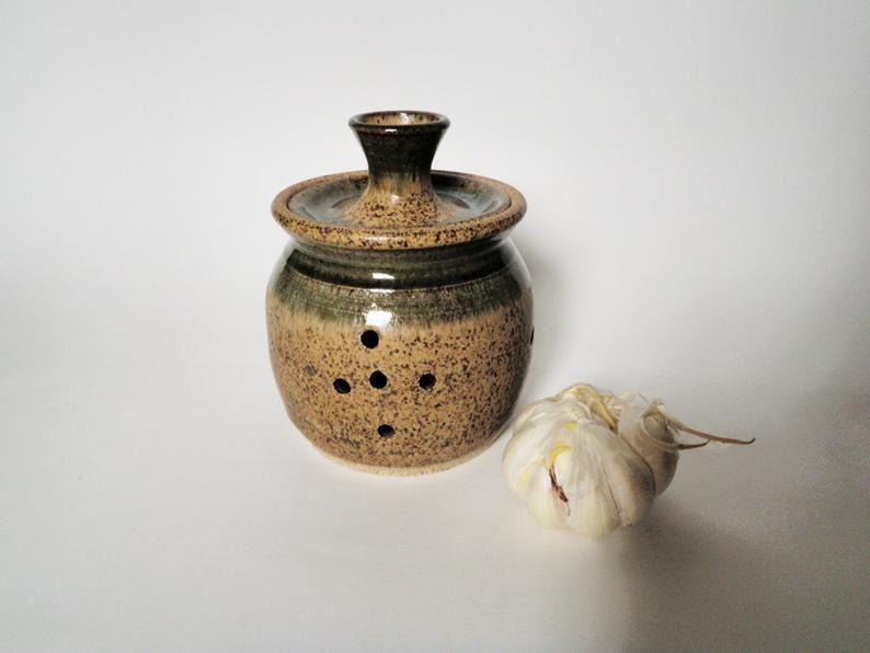 May include: A brown and green ceramic garlic keeper with a lid and holes for ventilation. A whole garlic bulb is sitting next to the keeper.