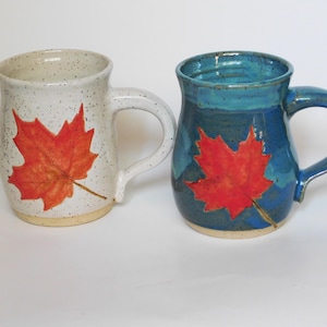 May include: Two ceramic mugs featuring a red maple leaf design. One mug is white with speckles and the other is blue.
