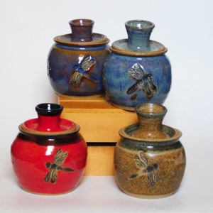 May include: Four ceramic jars with lids, each featuring a dragonfly design. The jars are in shades of blue, red, brown, and tan.