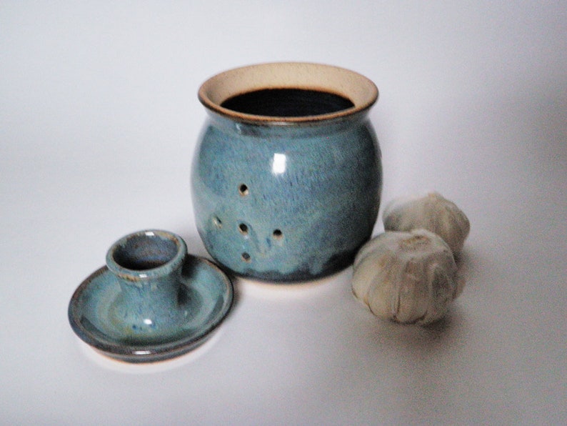 May include: A blue ceramic garlic keeper with a lid and a small dish for storing garlic cloves. The keeper has holes for air circulation and is glazed in a light blue color with brown accents.
