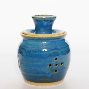 May include: A blue ceramic garlic keeper with a lid and holes for ventilation. The keeper has a light blue glaze with a subtle yellow hue.