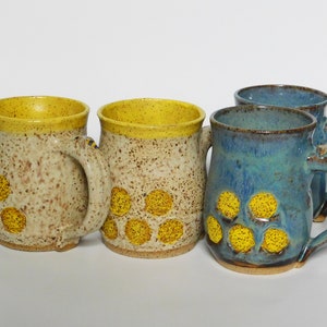May include: Four handmade ceramic mugs. Two are speckled beige with yellow rims and yellow dot patterns. The other two are blue with yellow dot patterns. All have large handles and are on a white background.