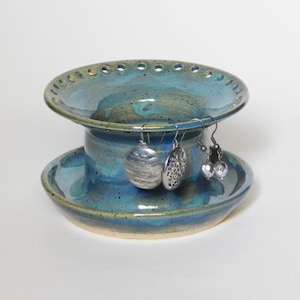 May include: A blue and green ceramic earring holder with a tiered design. The top tier has a wide, shallow bowl with holes around the edge. The bottom tier is a smaller, deeper bowl. Three silver earrings are hanging from the top tier.