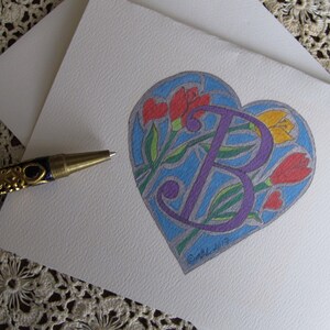 May include: A purple letter "B" surrounded by colorful flowers in a heart shape. The heart is outlined in black and has a blue background. The flowers are red, yellow, and green.