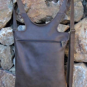 Könnte beinhalten: Ein brauner Lederrucksack mit verstellbaren Trägern und einer Reißverschlusstasche. Der Rucksack hängt an einer Steinmauer.