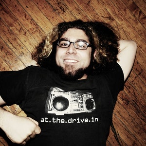 Può includere: Un uomo con i capelli castani e la barba indossa occhiali e una maglietta nera con una grafica bianca di un boombox e il testo "at.the.drive.in".