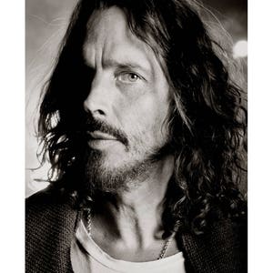 Op de afbeelding: Zwart-wit portretfoto van Chris Cornell, een muzikant, met lang haar en een baard. De foto is gemaakt in Seattle in 2012. De handtekening "Chris Cornell Seattle 2012" staat in de linkerbenedenhoek van de foto. De handtekening "Josh Baulkis" staat in de rechterbenedenhoek van de foto.