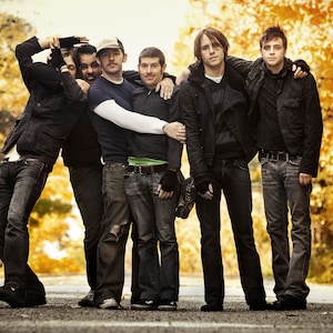 May include: Five men in a band pose for a photo in front of a fall foliage background. They are all wearing casual clothing, including jeans and jackets. The men are smiling and looking at the camera.