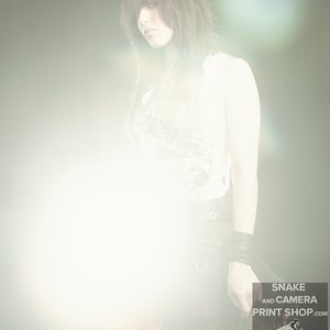 May include: A woman with dark hair and a white tank top stands in a bright light. She is wearing a black skirt with a red lace trim and fishnet stockings. The text "SNAKE AND CAMERA PRINT SHOP.COM" is visible in the lower right corner of the image.