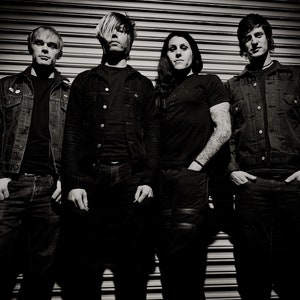 May include: A black and white photo of four people standing in front of a metal garage door. They are all wearing jeans and jackets. The person on the left is wearing a black jacket and the person on the right is wearing a blue jacket.