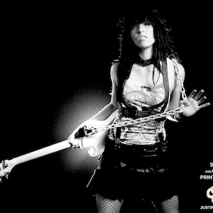 May include: A black and white photo of a woman with long black hair wearing a black choker necklace, a white tank top with a chain around her waist, a black skirt, and fishnet stockings. She is holding a white electric guitar and has a serious expression on her face. The background is black.
