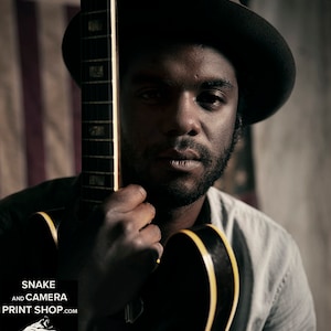 May include: A man wearing a black fedora and a grey shirt holds a black electric guitar with a yellow trim. The guitar is partially obscured by the man's body. The image includes the text "SNAKE AND CAMERA PRINT SHOP.COM" and "JUSTIN BORUCKI.COM".