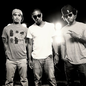 Puede incluir: Tres hombres con sombreros y gafas de sol se paran frente a un fondo negro con luces blancas. El hombre de la izquierda lleva una camiseta gris con un estampado gráfico en blanco y negro, pantalones caqui y una gorra de béisbol gris. El hombre del medio lleva una camiseta blanca, pantalones vaqueros azules y gafas de sol negras. El hombre de la derecha lleva una camiseta gris, pantalones vaqueros azules, una gorra de béisbol negra y gafas de sol negras.