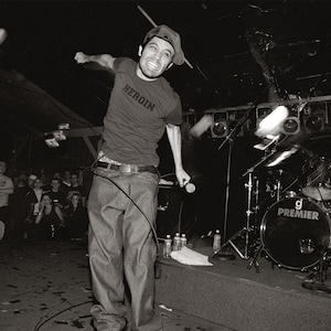 May include: A black and white photo of a band performing on stage. The lead singer is wearing a black shirt with the word "HEROIN" printed on it. He is holding a microphone and singing into it. The drummer is behind him, playing a drum set with a "Premier" logo. The band is performing in front of a crowd of people.