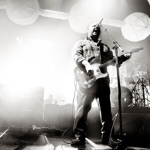 May include: A man in a plaid shirt sings into a microphone while playing a black electric guitar on stage. He is performing in front of a crowd of people.