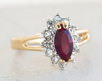 Puede incluir: Un anillo de oro con una gran piedra roja rodeada de piedras pequeñas transparentes.