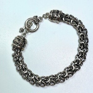 Puede incluir: Una pulsera plateada con cierre de palanca. La pulsera est&aacute; hecha de eslabones redondeados interconectados, creando un efecto de cota de malla. El cierre y los elementos decorativos presentan detalles intrincados. La joya se exhibe sobre una superficie blanca.