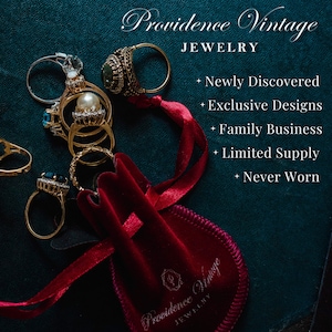 May include: A red velvet pouch with the text "Providence Vintage Jewelry" on it, surrounded by a red ribbon and a collection of vintage rings with gold and silver settings, including a pearl ring and a ring with a blue gemstone.