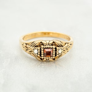 May include: A gold ring featuring a square red gemstone surrounded by smaller clear gemstones. The ring has an intricate floral design.