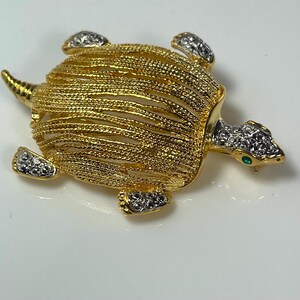 May include: A gold-toned turtle brooch with a textured shell design. The turtle's head and feet are adorned with silver-colored rhinestones. A small green gem accents the eye.