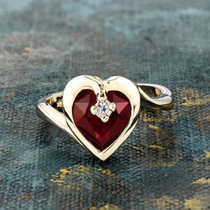 May include: A gold heart-shaped ring with a red gemstone and a small diamond accent.