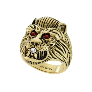Può includere: Un anello in oro a forma di testa di leone con occhi di pietre preziose rosse e un solo diamante in bocca. L'anello è contrassegnato "18KGE".