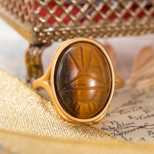 May include: A gold-toned ring featuring an oval-shaped tiger's eye gemstone. The stone is carved with a sunburst design, displaying brown and golden chatoyant bands. The metal band has a smooth, polished finish and a split-shank design.