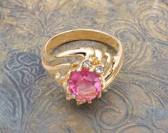May include: A gold ring with a pink oval-shaped gemstone and two smaller clear gemstones. The ring is on a gold-colored background.